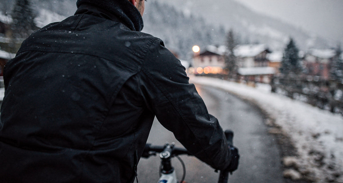 Blick über die Schulter eines Radfahrers in Winterkleidung auf eine verschneite Alpenstraße im Schneetreiben